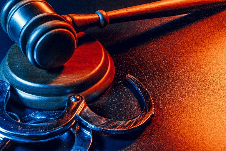 An up close dramatically lit orange and blue image shows a gavel and pair of handcuffs. 